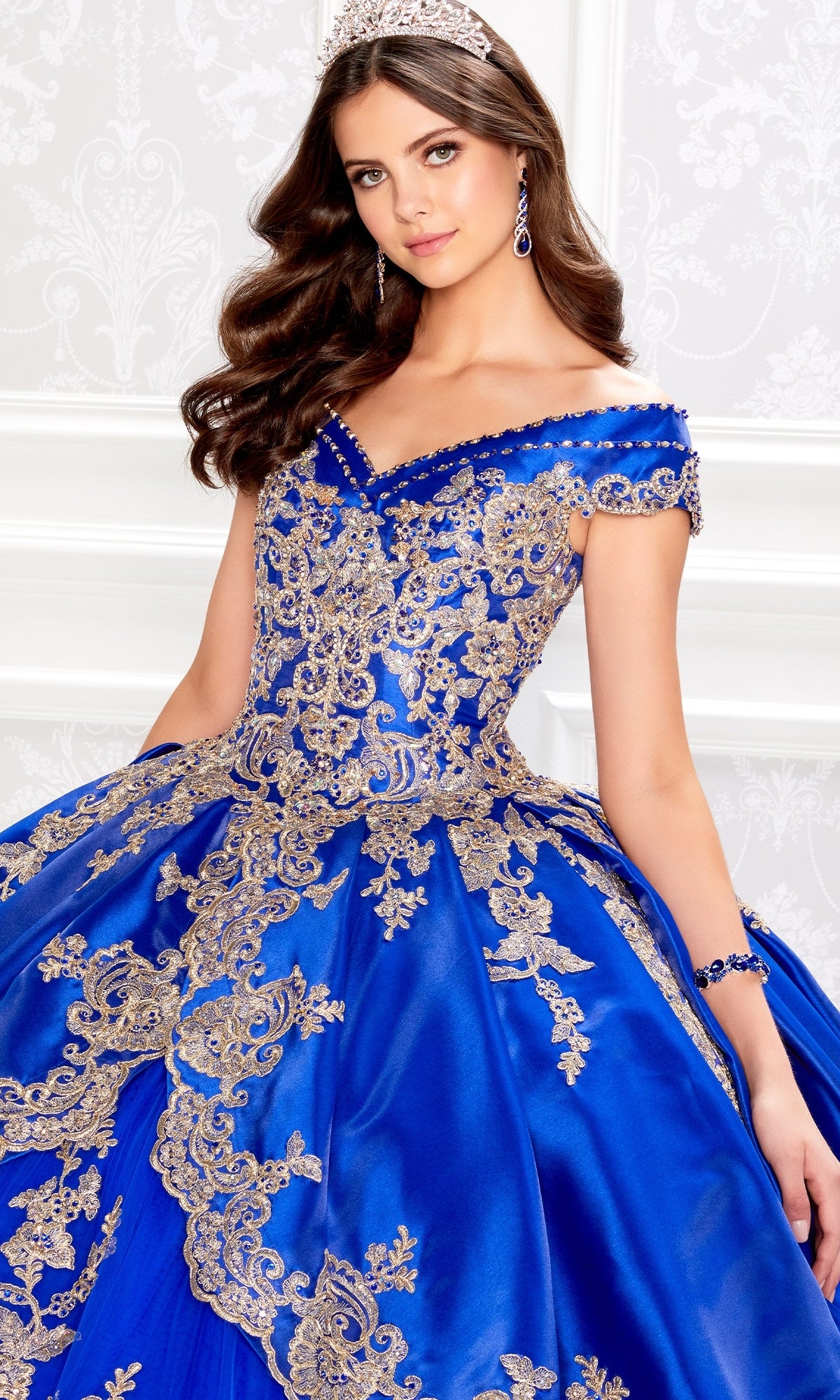 A woman stands in a white ornate room wearing the Princesa PR22029 satin quinceanera dress, featuring off-the-shoulder royal blue fabric with gold embroidery, paired with matching accessories and a tiara.