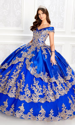 A young woman in the Princesa PR22029 Satin Quinceanera Dress, featuring off-the-shoulder blue satin with gold embroidery and a tiara, stands before a white decorative wall.