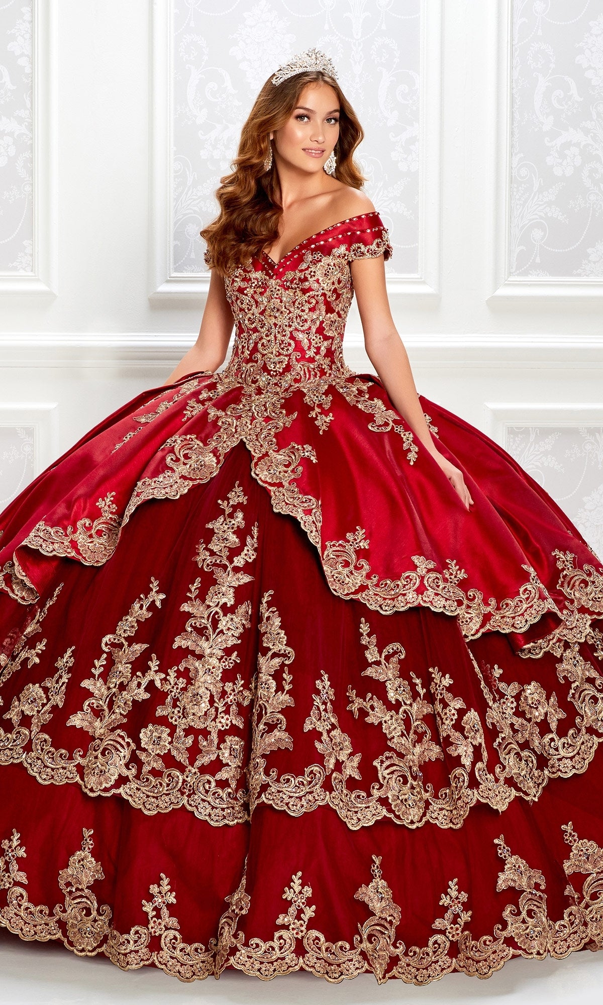 A woman poses against a decorative white wall in the Princesa PR22029 Satin Quinceanera Dress, an ornate off-the-shoulder red gown with gold embroidery and a tiara.