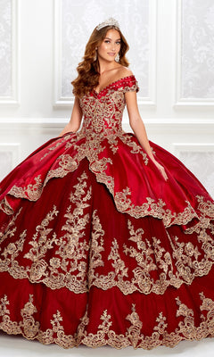 A woman poses against a decorative white wall in the Princesa PR22029 Satin Quinceanera Dress, an ornate off-the-shoulder red gown with gold embroidery and a tiara.