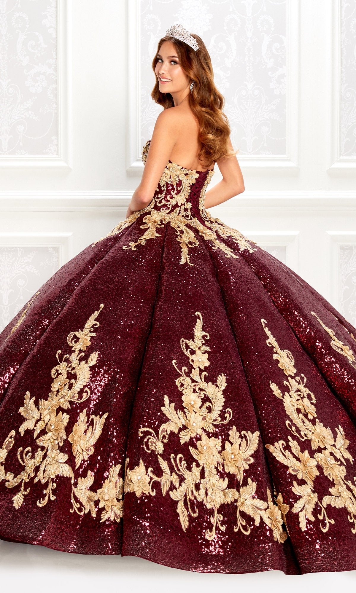 A woman in the Wine Red Quinceanera Dress: Princesa PR22030, featuring gold embroidery and sequins, stands sideways wearing a tiara before an ornate white wall.