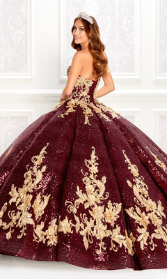 A woman in the Wine Red Quinceanera Dress: Princesa PR22030, featuring gold embroidery and sequins, stands sideways wearing a tiara before an ornate white wall.