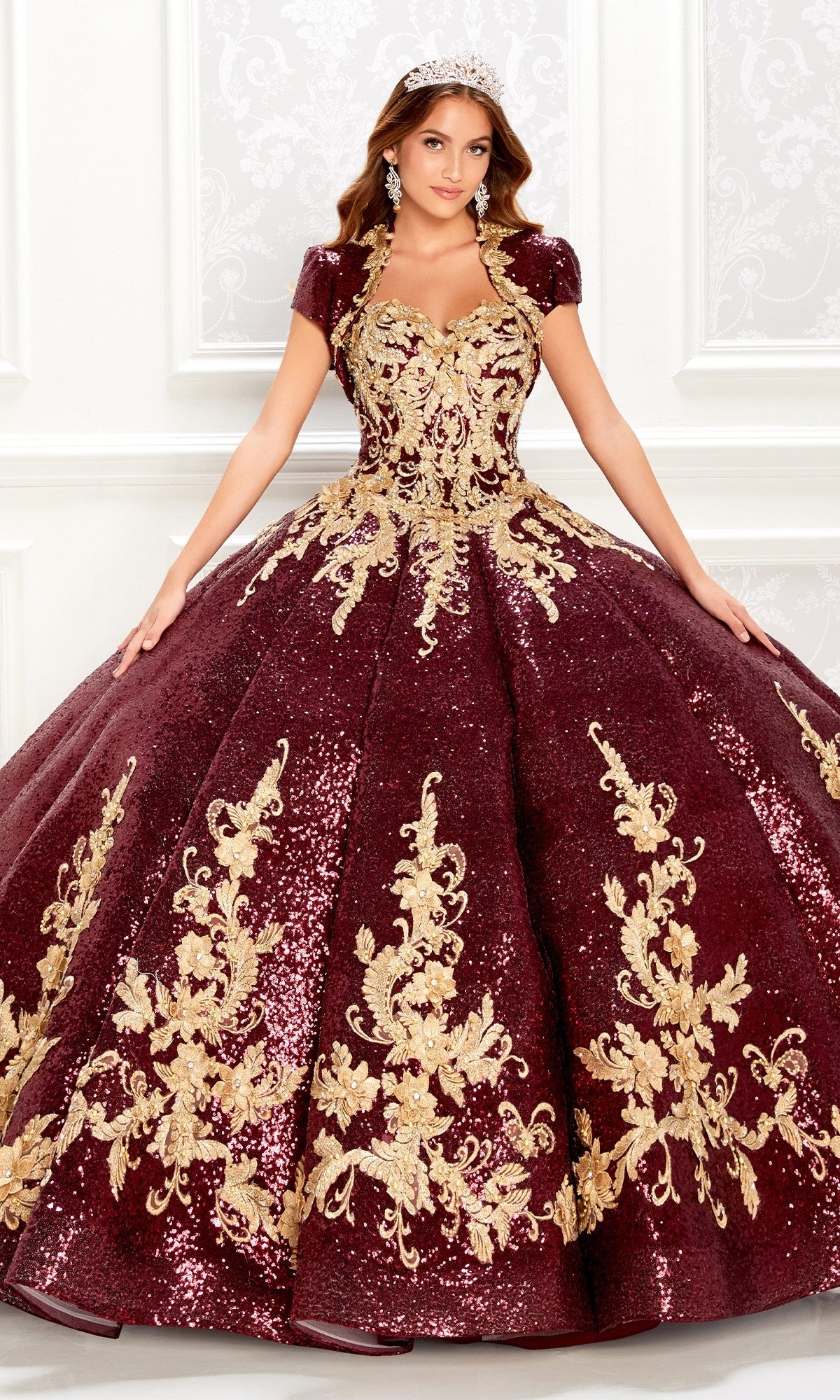 A woman in the Princesa PR22030 Wine Red Quinceanera Dress with gold embroidery and sequins, wearing a tiara, stands before an ornate white wall.