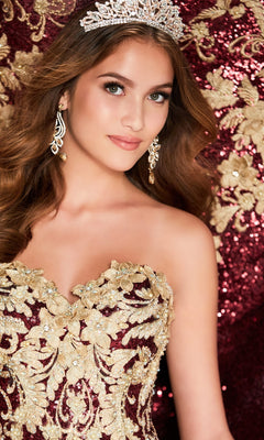 A woman wearing a jeweled tiara, gold earrings, and the Wine Red Quinceanera Dress: Princesa PR22030 with gold embroidery and lace appliques poses before a coordinating burgundy and gold floral backdrop.