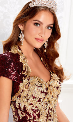 A woman wearing the Wine Red Quinceanera Dress: Princesa PR22030, adorned with gold embroidery and lace appliques, paired with a jeweled tiara and dangling earrings, poses and smiles slightly against a light-patterned background.