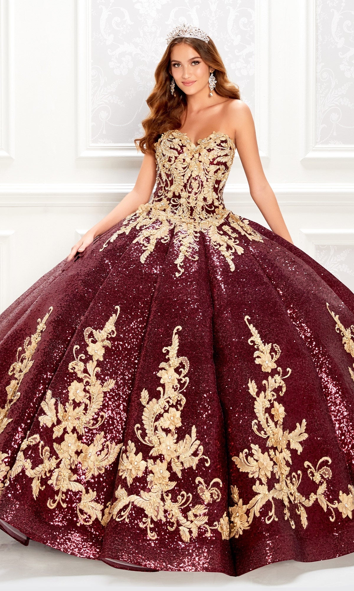 A woman poses indoors against a white decorated wall wearing the Princesa PR22030, a wine red quinceanera dress featuring ornate gold floral embroidery, sequins, and delicate lace appliques.