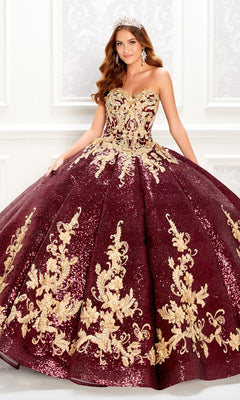 A woman poses indoors against a white decorated wall wearing the Princesa PR22030, a wine red quinceanera dress featuring ornate gold floral embroidery, sequins, and delicate lace appliques.