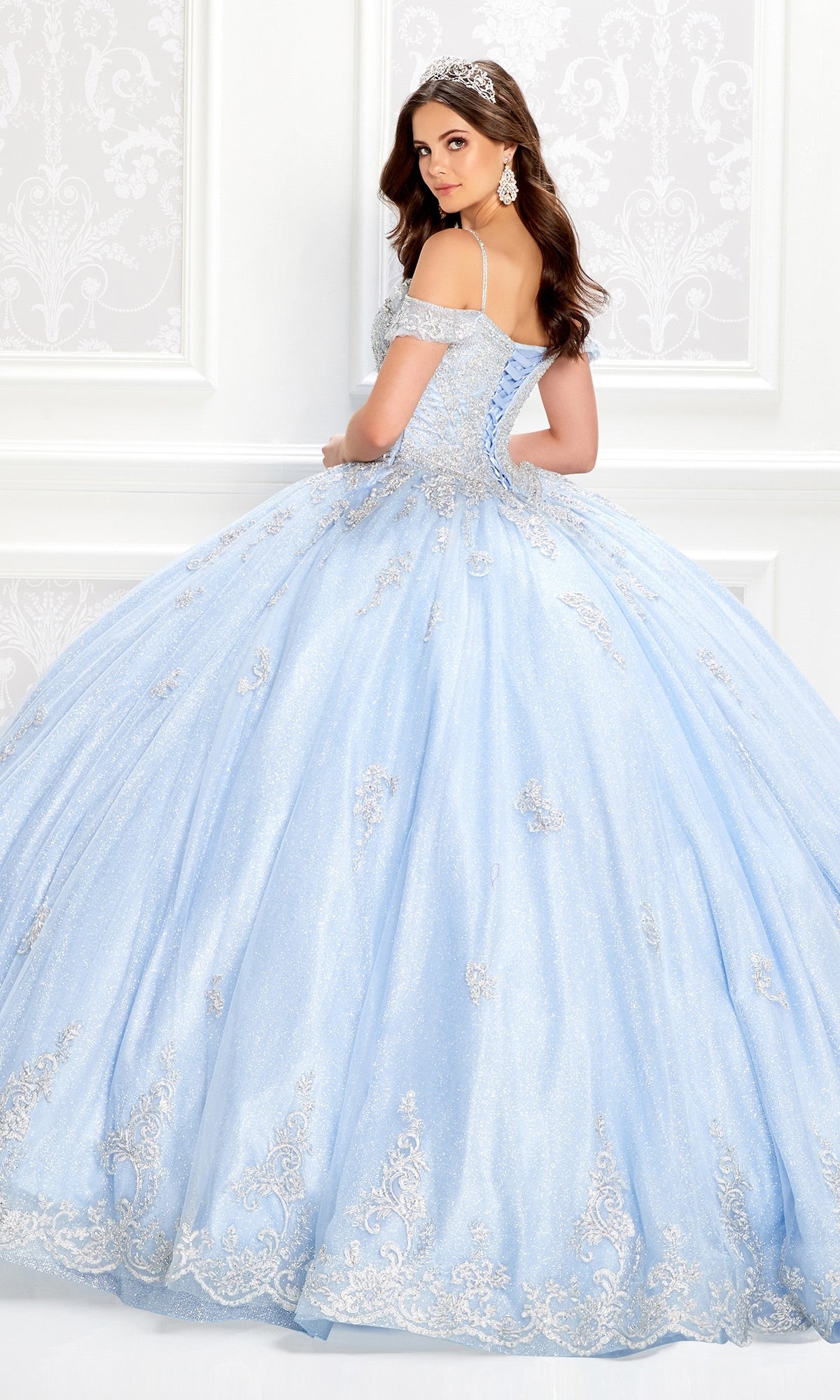A woman in the Light Blue Quinceanera Dress: Princesa PR22032 with lace appliques and a corset bodice glances over her shoulder in an ornate, light-colored room.