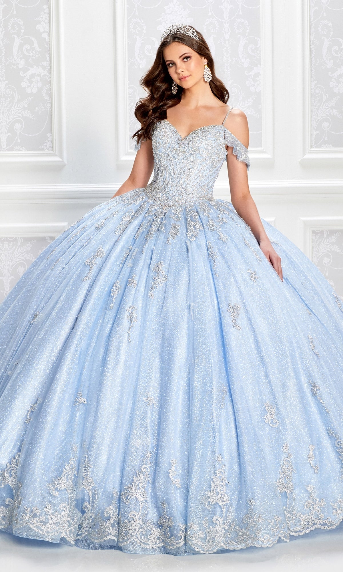 A woman in the Light Blue Quinceanera Dress: Princesa PR22032, with a corset bodice and silver accents, stands in an ornate white room as her tiara sparkles under the lights.