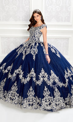 A woman in the Quinceanera Dress PR22035 By Princesa, a navy blue gown with silver embroidery and a corset bodice, stands before a white ornate wall, holding out her skirt.