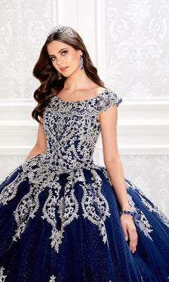 A young woman in the PR22035 Quinceanera Dress by Princesa, featuring silver embroidery, glitter tulle, and a corset bodice, stands against an ornate white background wearing a jeweled tiara.