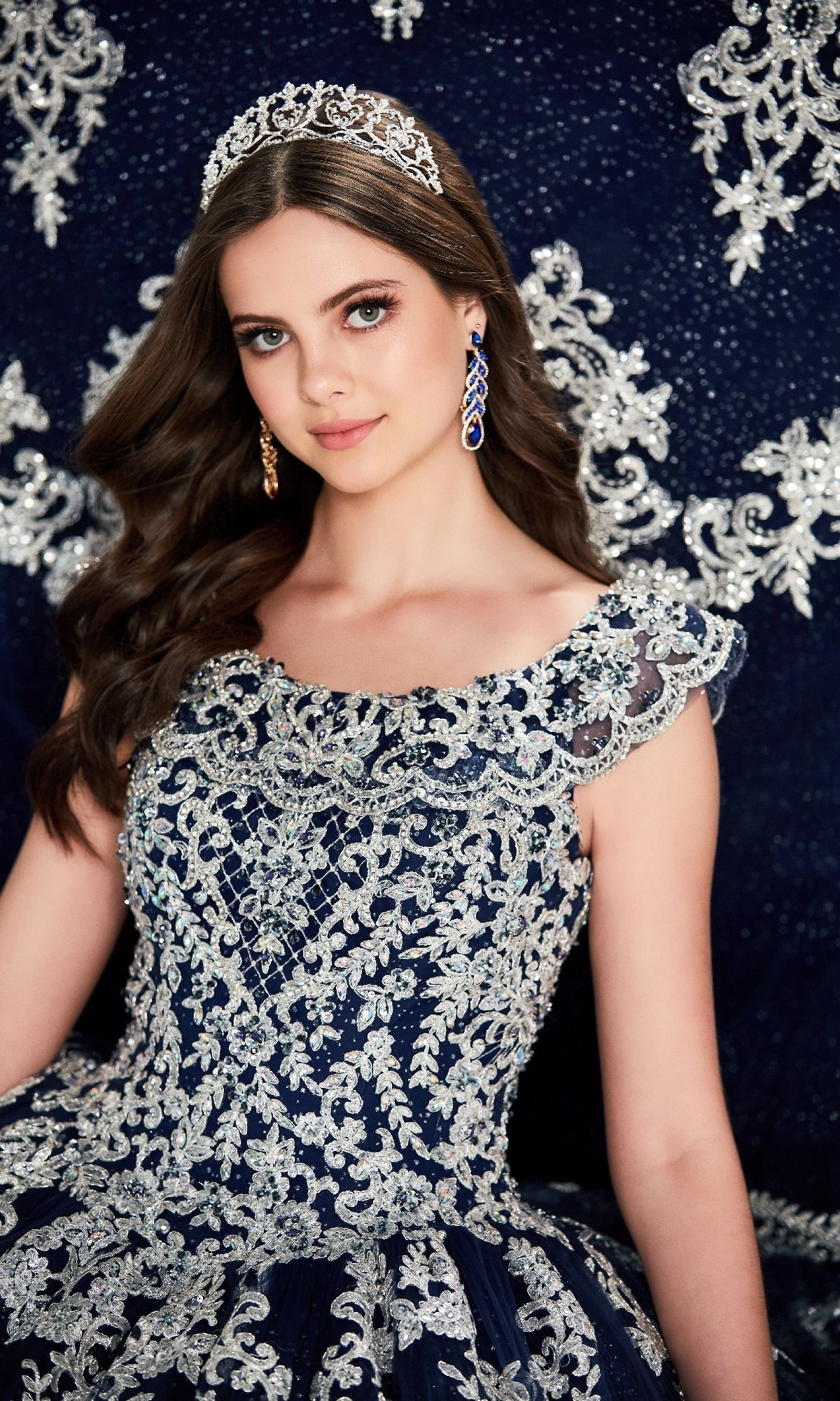 A young woman wears the Quinceanera Dress PR22035 By Princesa in navy blue with silver embroidery and a corset bodice, paired with a tiara and matching earrings, as she poses before a coordinating patterned background.