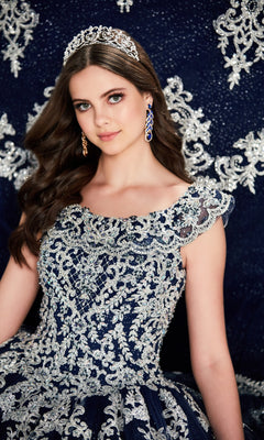 A young woman wears the Quinceanera Dress PR22035 By Princesa in navy blue with silver embroidery and a corset bodice, paired with a tiara and matching earrings, as she poses before a coordinating patterned background.
