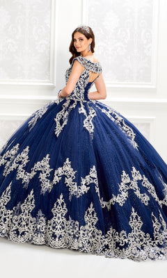Woman wears the Quinceanera Dress PR22035 By Princesa, a dark blue ball gown with a glitter tulle skirt and silver embroidery, posing in an elegant white room while looking over her shoulder and wearing a jeweled headband.