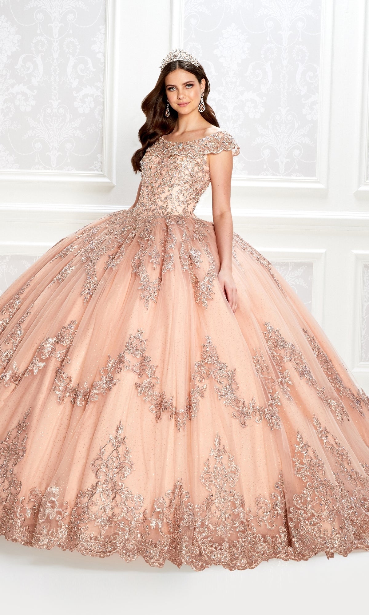 A young woman wears the Quinceanera Dress PR22035 By Princesa, featuring peach glitter tulle and silver embellishments, with a tiara, as she stands in an ornately decorated room with white patterned walls.