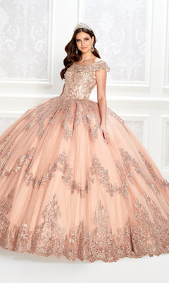 A young woman wears the Quinceanera Dress PR22035 By Princesa, featuring peach glitter tulle and silver embellishments, with a tiara, as she stands in an ornately decorated room with white patterned walls.