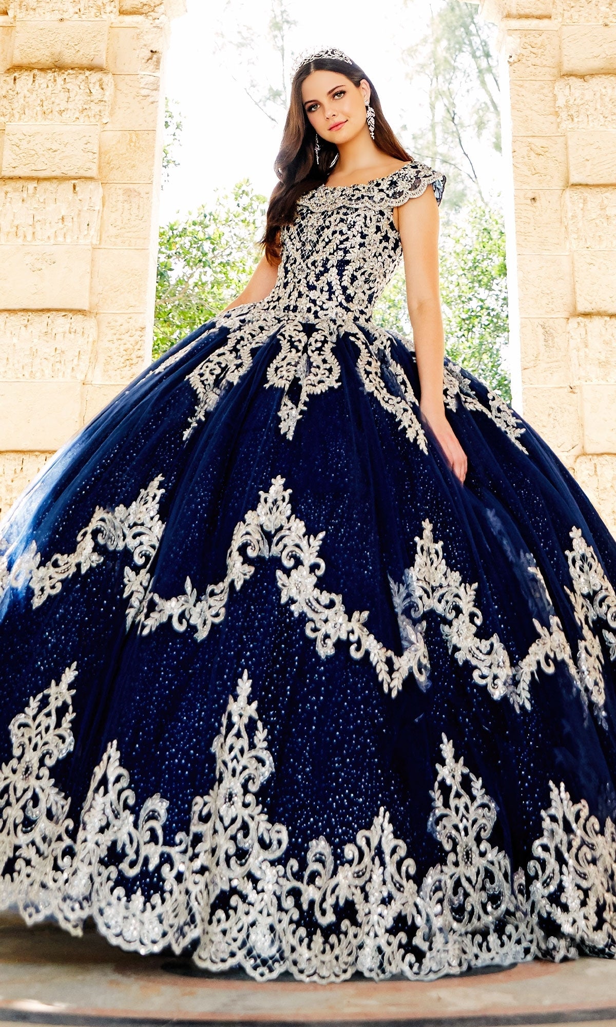 A woman stands outdoors in front of a stone archway, wearing the Quinceanera Dress PR22035 by Princesa—a dark blue gown with ornate silver embroidery and a tiara.