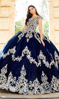 A woman stands outdoors in front of a stone archway, wearing the Quinceanera Dress PR22035 by Princesa—a dark blue gown with ornate silver embroidery and a tiara.