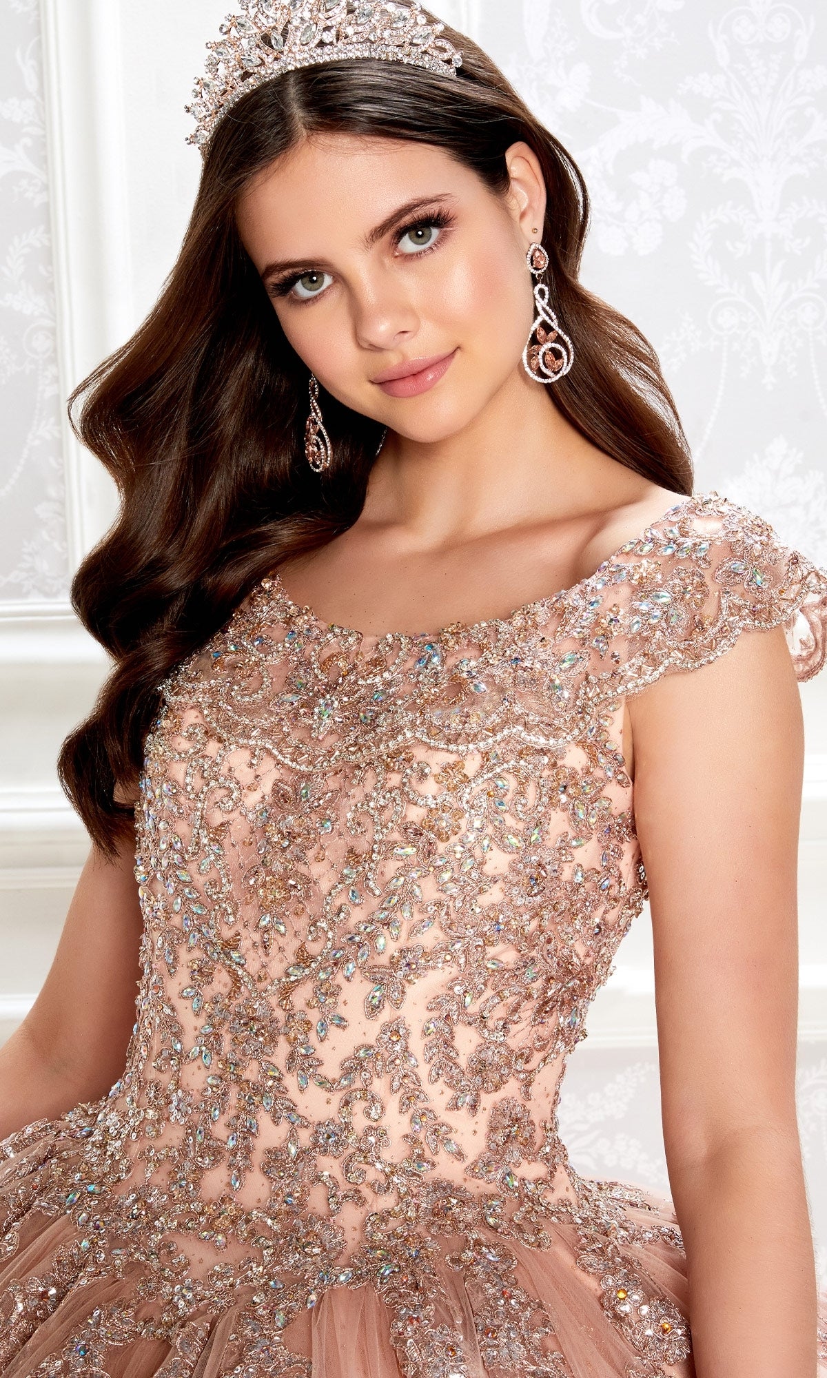 A young woman wearing a jeweled tiara, dangling earrings, and the Quinceanera Dress PR22035 By Princesa with a glitter tulle, off-the-shoulder design and embellished corset bodice poses against a light patterned background.