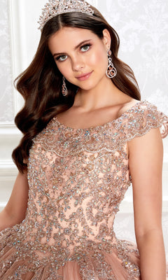 A young woman wearing a jeweled tiara, dangling earrings, and the Quinceanera Dress PR22035 By Princesa with a glitter tulle, off-the-shoulder design and embellished corset bodice poses against a light patterned background.
