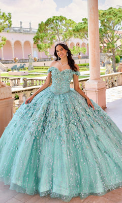 A young woman smiles on an outdoor terrace, wearing the Flower Quince Dress: Princesa by Ariana Vara PR22036—a sparkling light blue ball gown with a tiara, framed by columns and garden views.