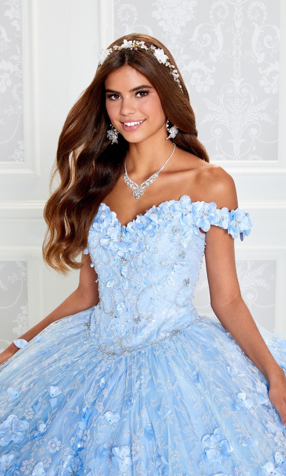 Young woman in the Flower Quince Dress: Princesa by Ariana Vara PR22036, a blue off-the-shoulder ball gown with floral details, accessorized with a tiara, necklace, and earrings, posing before a white patterned wall.