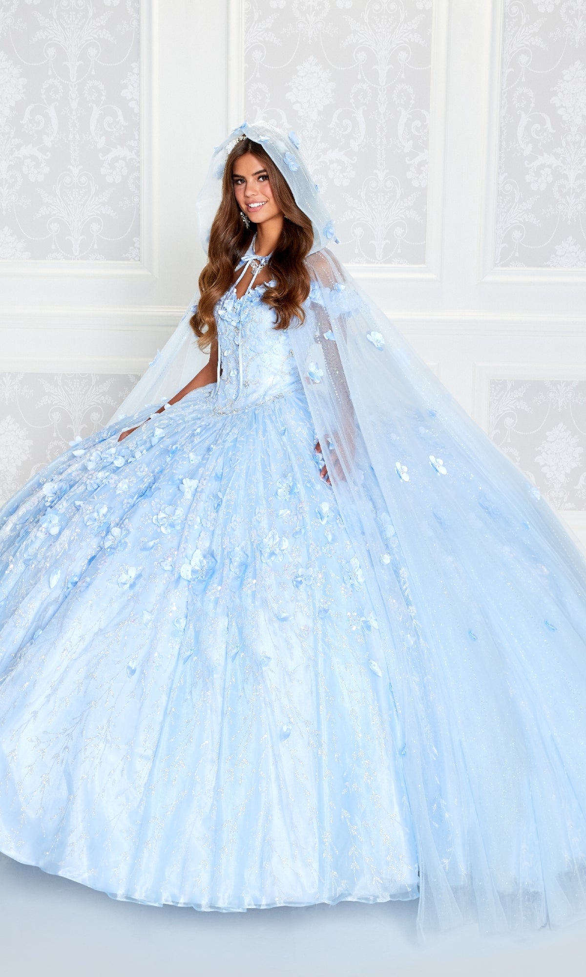 A young woman in the Flower Quince Dress: Princesa by Ariana Vara PR22036—with light blue floral details and a matching cape—stands before a white patterned wall, radiating dreamy elegance.