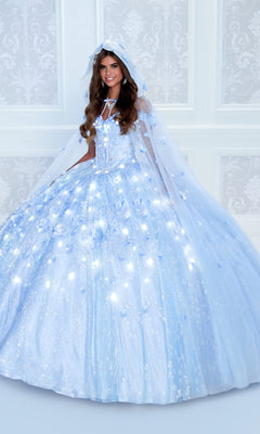 A woman stands in front of a white decorative wall wearing the Flower Quince Dress: Princesa by Ariana Vara PR22036, a light blue glow-in-the-dark quinceañera gown with a hood and floral details.