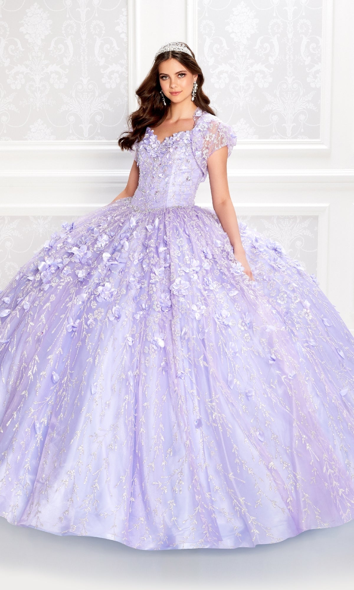 A young woman wears the Flower Quince Dress: Princesa by Ariana Vara PR22036—a light purple ball gown with floral details and a tiara—standing before a white patterned wall.