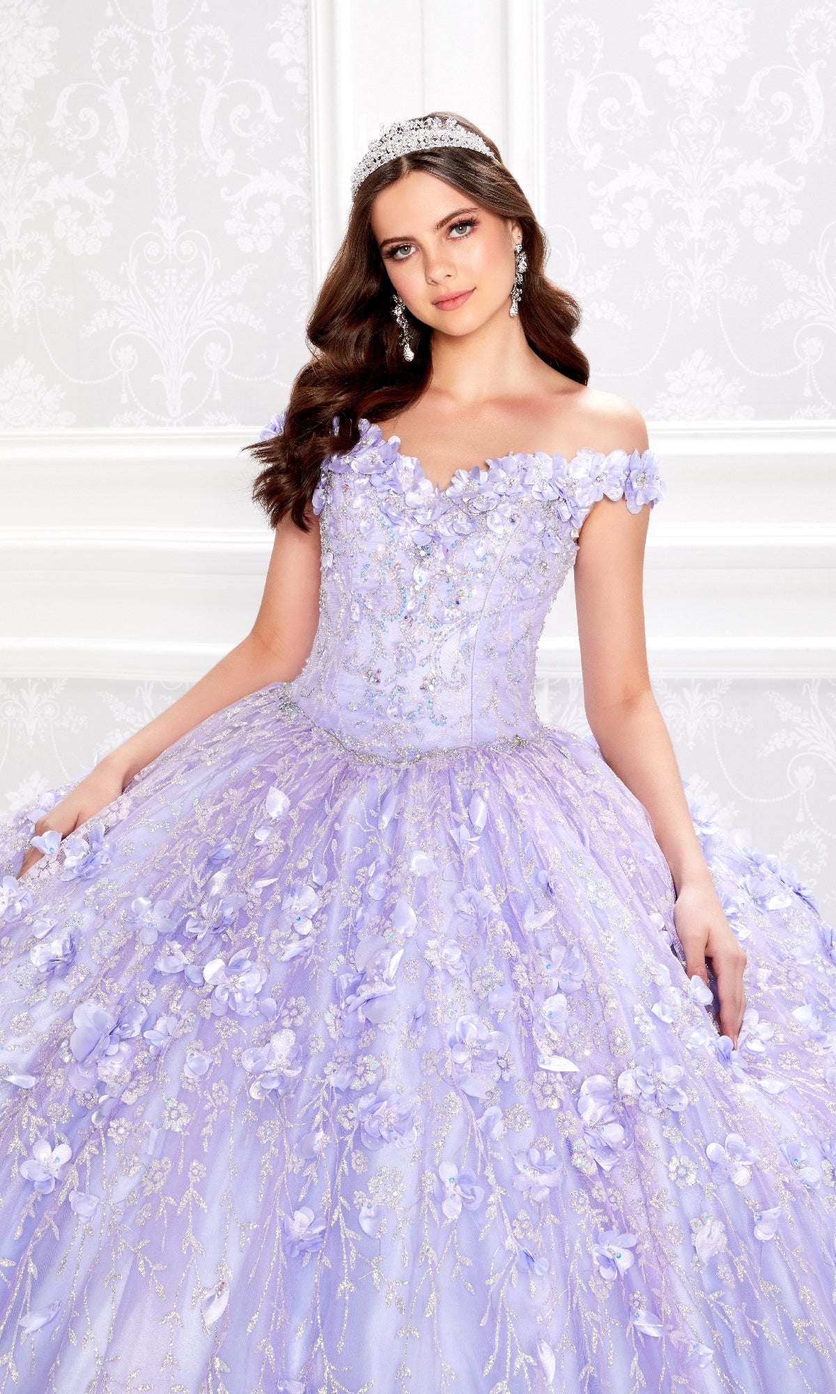 A young woman poses in front of a white ornate wall wearing the Flower Quince Dress: Princesa by Ariana Vara PR22036, a lavender off-shoulder ball gown with floral embellishments and a tiara.