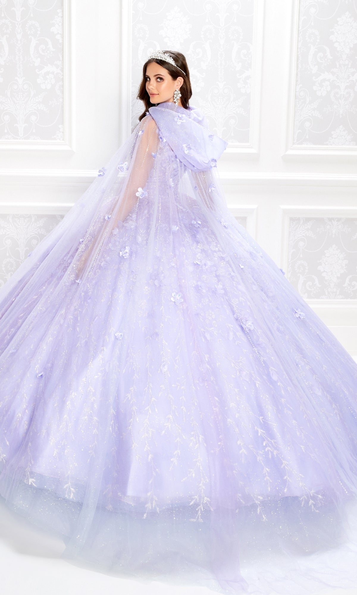 A woman wearing the Flower Quince Dress: Princesa by Ariana Vara PR22036, a light purple ballgown with a long cape, stands before a white ornate wall and looks over her shoulder.