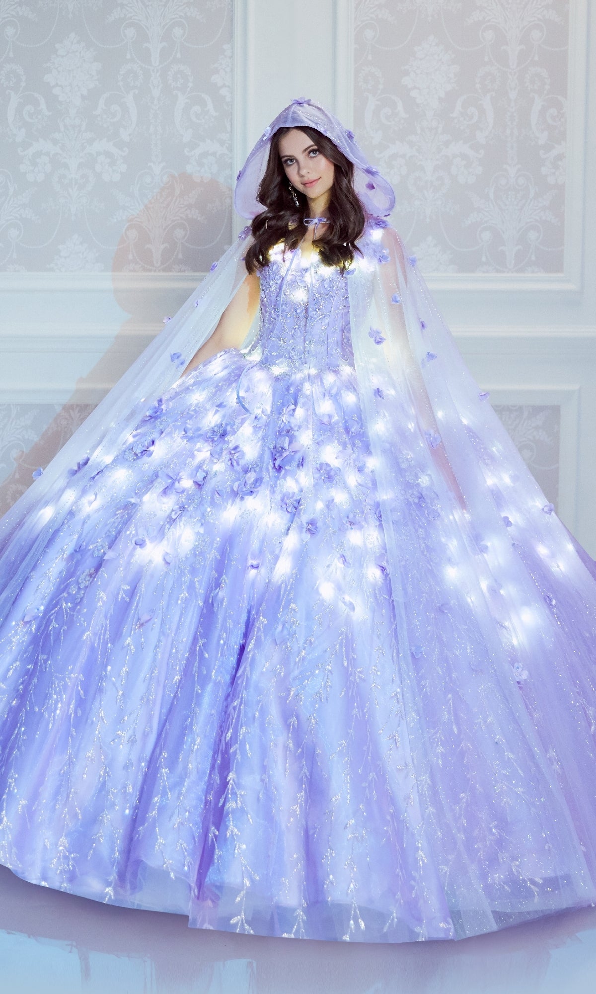 A woman stands in a white room wearing the Princesa by Ariana Vara PR22036 Flower Quince Dress, a lavender ball gown with a matching hooded cape illuminated by small white lights for a magical glow-in-the-dark effect.