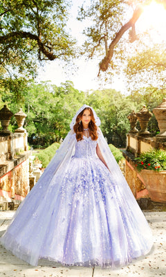 A young woman in the Flower Quince Dress: Princesa by Ariana Vara PR22036, a light purple ball gown with matching cape and hood, stands outdoors on stone steps surrounded by greenery and sunlight.