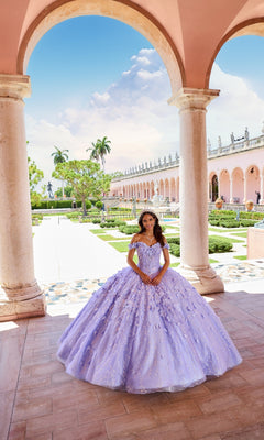 A young woman in a lavender Flower Quince Dress: Princesa by Ariana Vara PR22036 stands under an archway in a formal garden with palm trees and a pink building in the background.