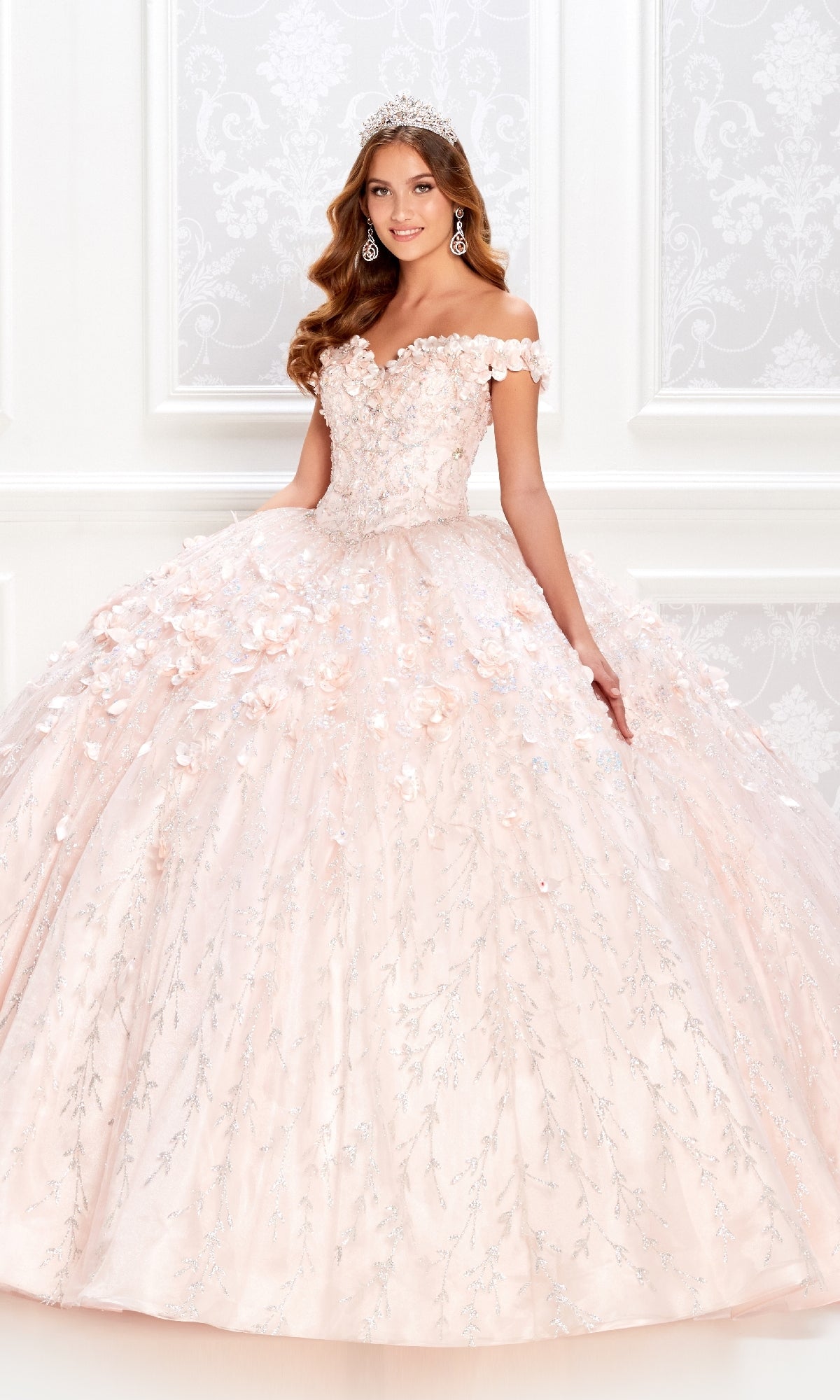 A young woman wearing the Flower Quince Dress: Princesa by Ariana Vara PR22036—a pink, off-the-shoulder ball gown with floral appliqués and a tiara—stands indoors before a patterned white wall.