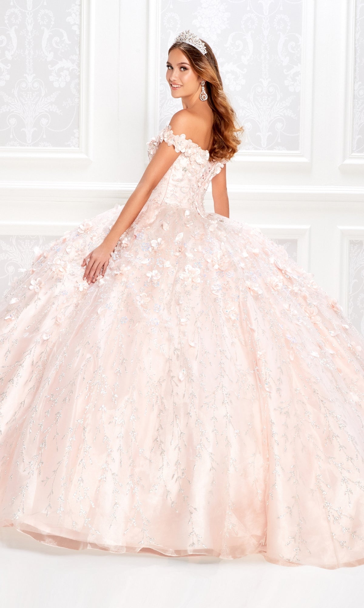 A young woman in the Princesa by Ariana Vara PR22036 Flower Quince Dress, wearing a tiara, stands in an elegant white-paneled room, smiling as she looks over her shoulder.
