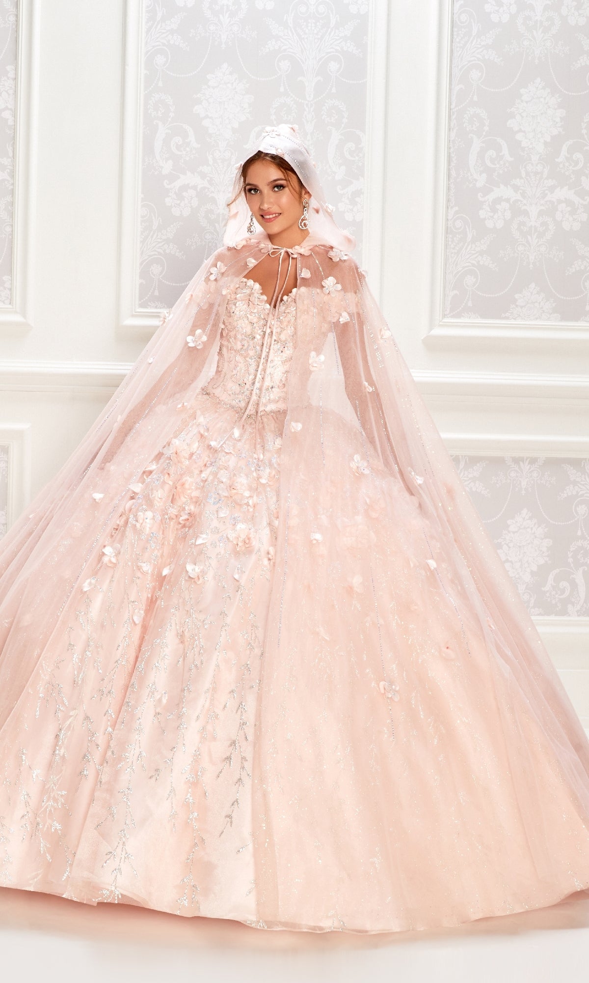 A woman in a light pink Flower Quince Dress: Princesa by Ariana Vara PR22036, featuring embellishments and a matching hooded cape, stands before a white patterned wall.
