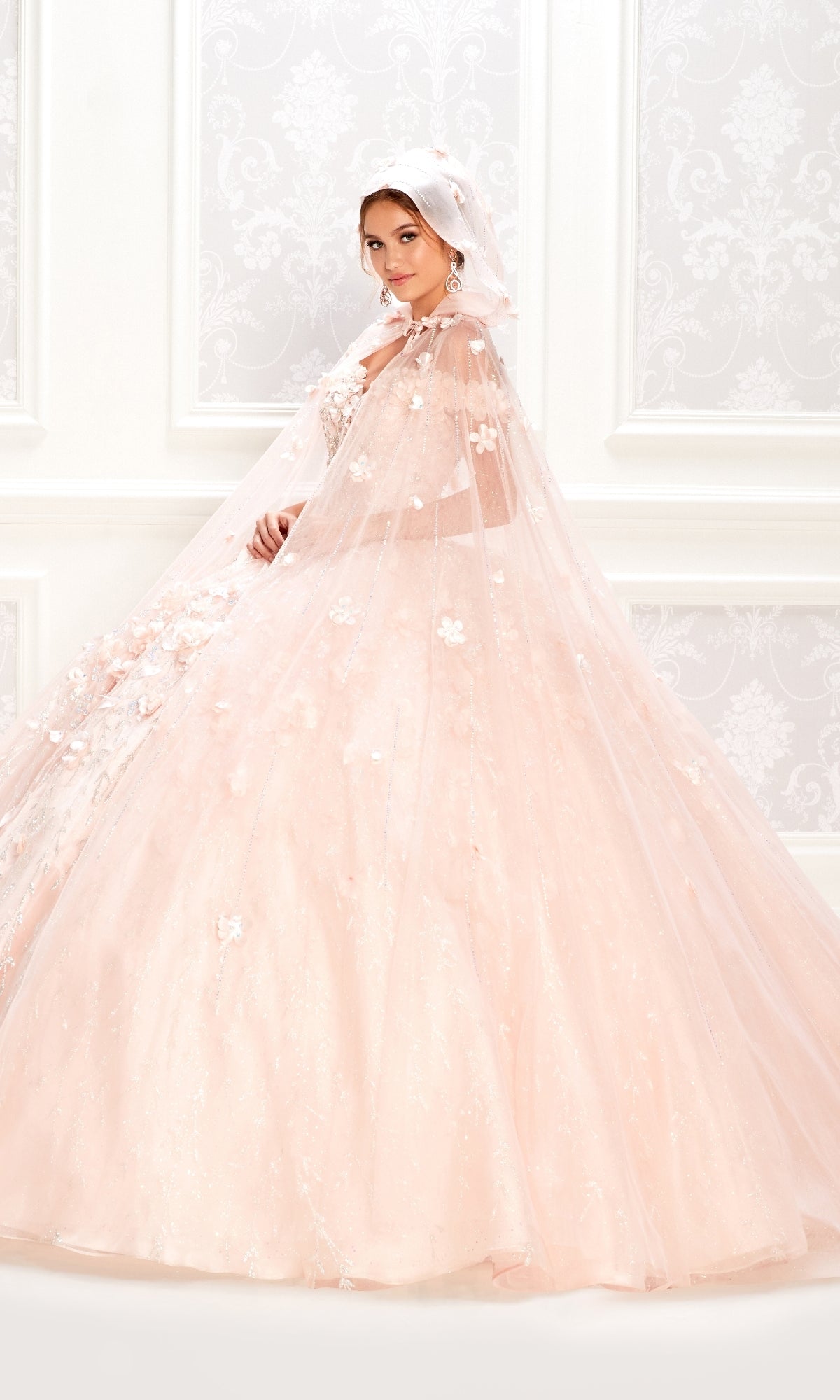 A woman in the Flower Quince Dress (Princesa by Ariana Vara PR22036), a light pink embellished ball gown with matching cape and headscarf, stands before a white ornate background.