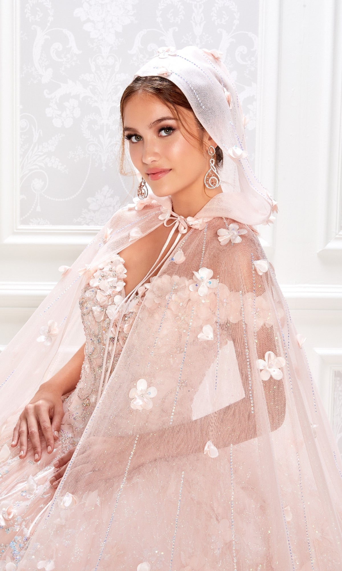 A woman poses indoors against an ornate white background, wearing the Flower Quince Dress: Princesa by Ariana Vara PR22036 in light pink, adorned with floral embellishments and a sheer hooded cape.