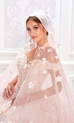 A woman poses indoors against an ornate white background, wearing the Flower Quince Dress: Princesa by Ariana Vara PR22036 in light pink, adorned with floral embellishments and a sheer hooded cape.
