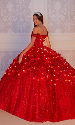 A young woman in the Flower Quince Dress: Princesa by Ariana Vara PR22036, a sparkling red ballgown with built-in lights, stands before an ornate wall, smiling over her shoulder and wearing a silver tiara.