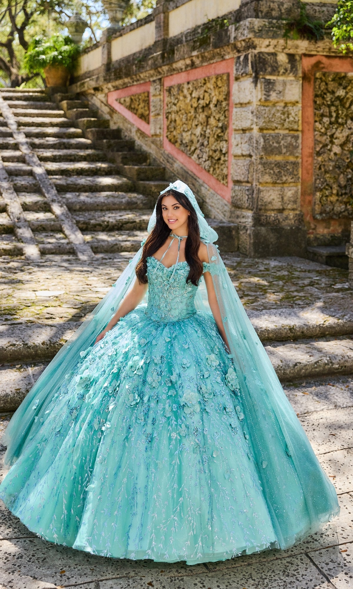 A young woman stands outdoors on stone steps, beside a textured wall and greenery, wearing the Princesa by Ariana Vara PR22036 Flower Quince Dress and a veil.