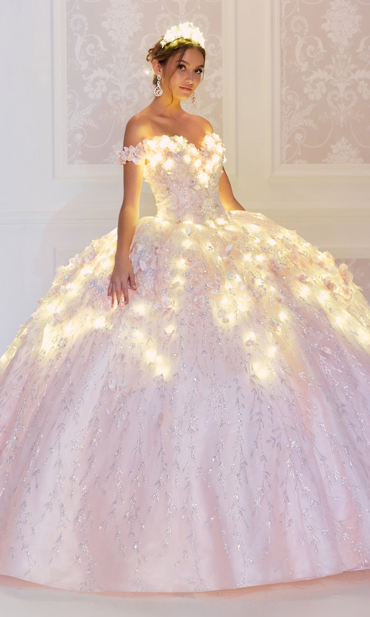 A young woman wears the Flower Quince Dress: Princesa by Ariana Vara PR22036, a light pink, off-the-shoulder ball gown with floral details and integrated lights, glowing softly as she stands against an ornate, light-colored wall.