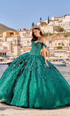 A woman in a green Flower Quince Dress: Princesa by Ariana Vara PR22036 and tiara stands by the water with colorful hillside buildings in the background.