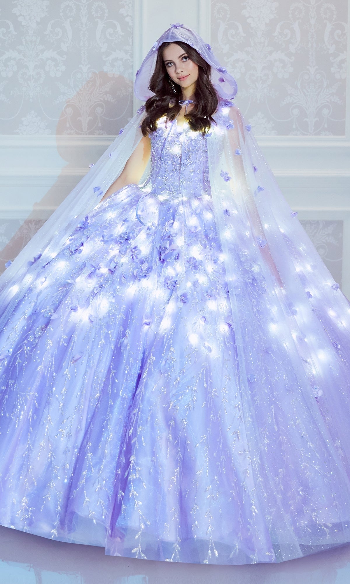 A woman in the Flower Quince Dress: Princesa by Ariana Vara PR22036, featuring light purple glow-in-the-dark fabric, floral accents, and a matching hooded cape, stands before a white patterned wall.