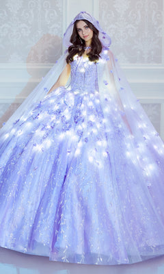 A woman in the Flower Quince Dress: Princesa by Ariana Vara PR22036, featuring light purple glow-in-the-dark fabric, floral accents, and a matching hooded cape, stands before a white patterned wall.