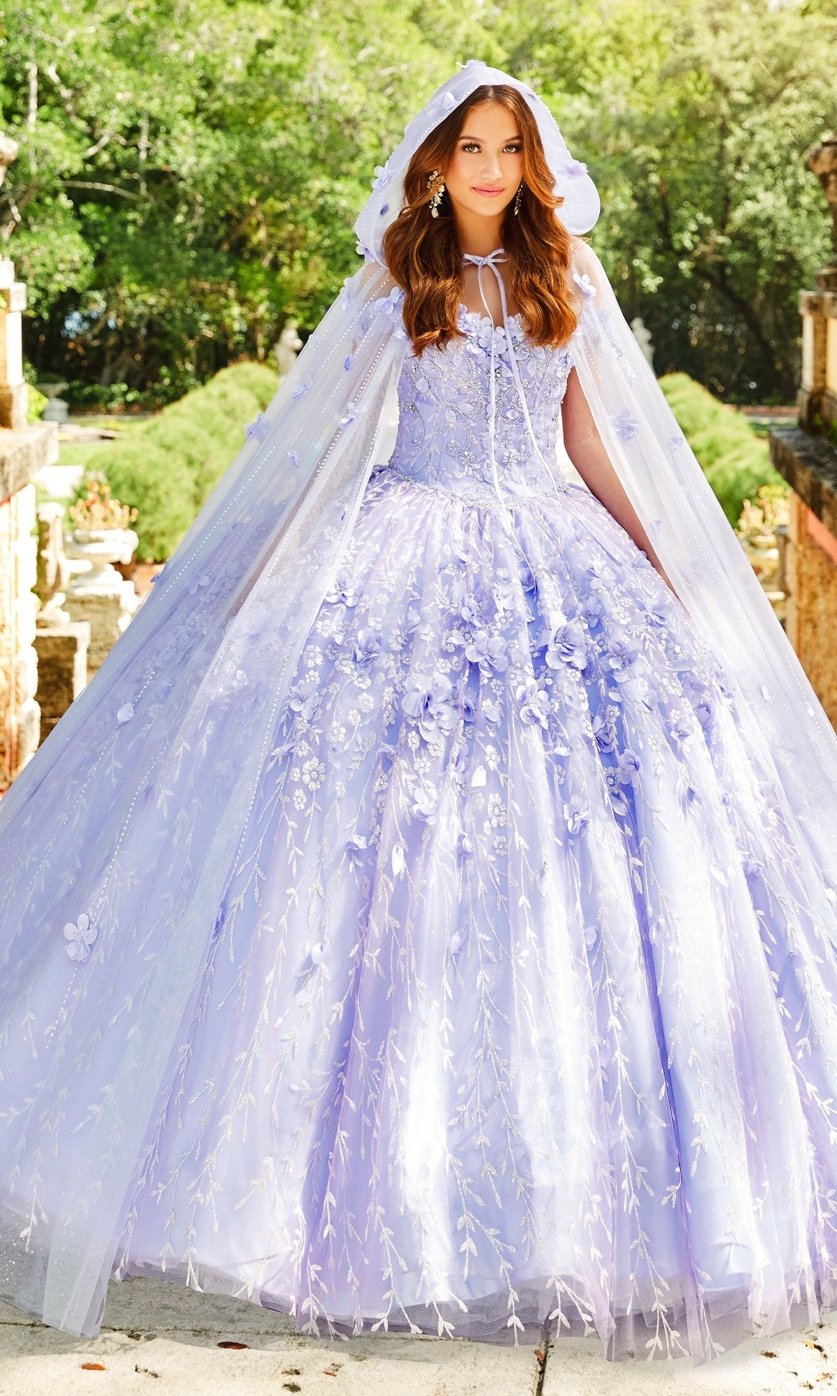 A woman stands outdoors wearing the Princesa by Ariana Vara PR22036 Flower Quince Dress in light purple with floral embroidery, paired with a matching hooded cape, surrounded by trees and stone structures.