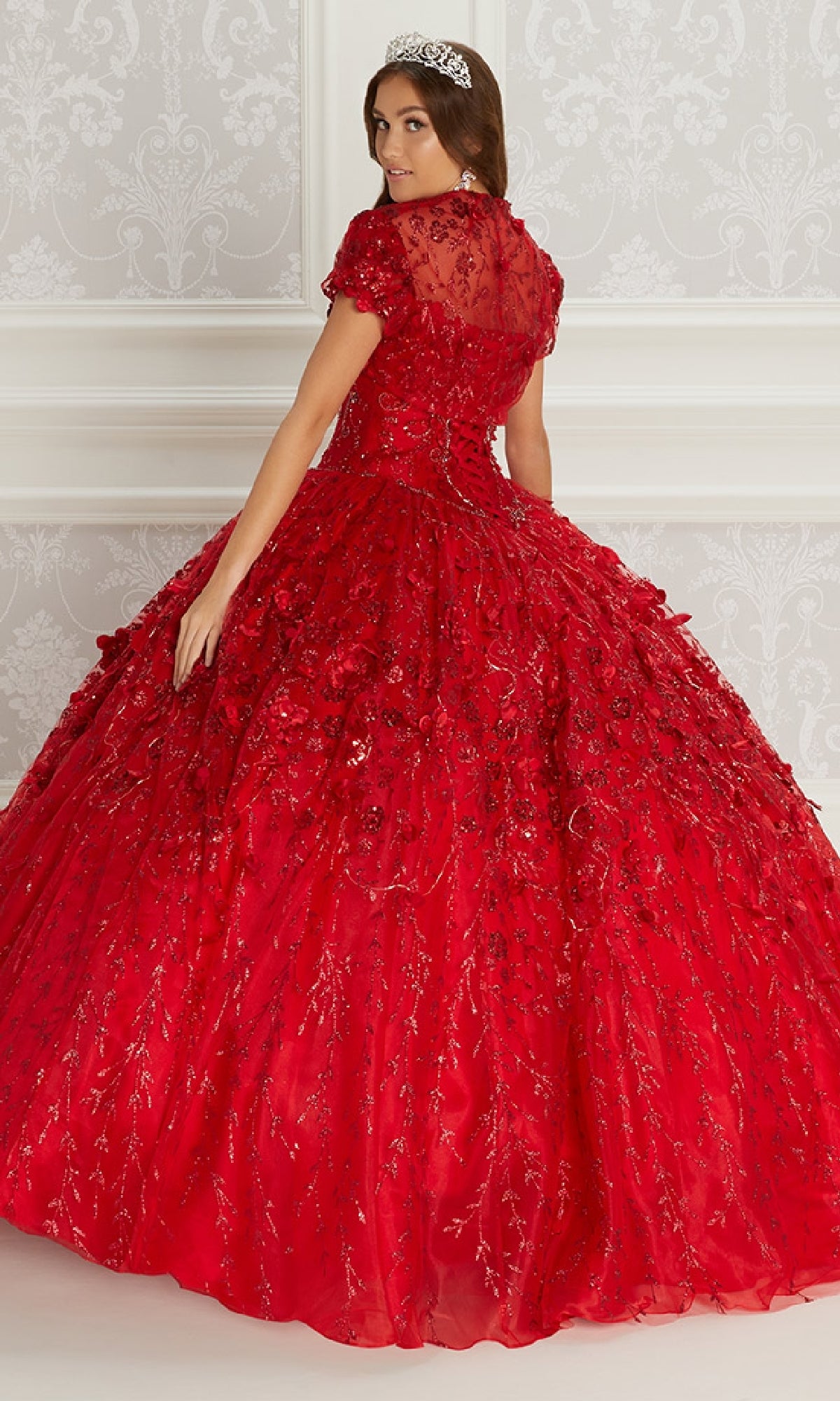 A young woman wears the Flower Quince Dress: Princesa by Ariana Vara PR22036, a voluminous red gown with jeweled tiara and embroidered floral details, posing before a white patterned wall.