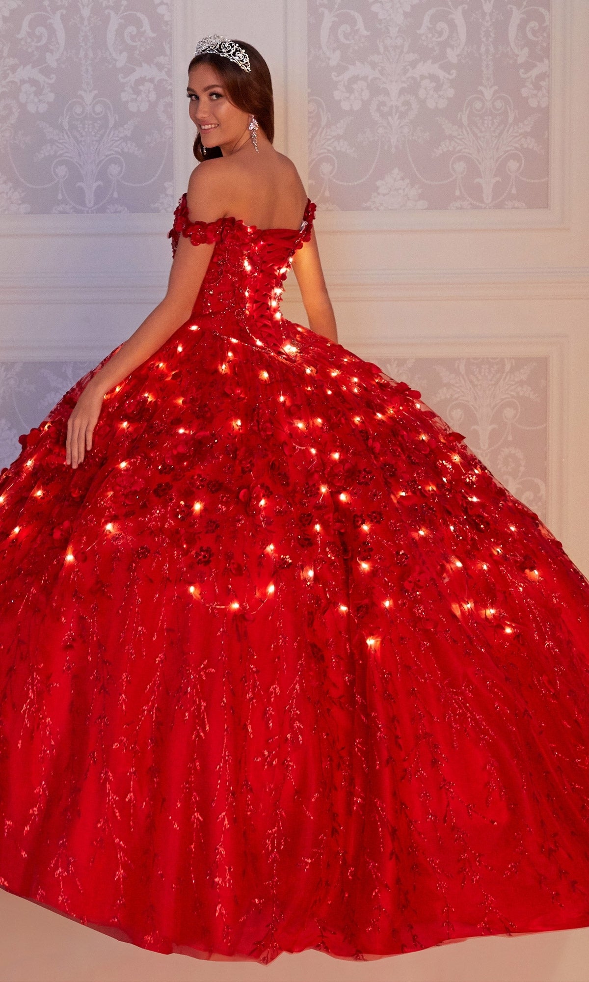 A young woman in the glow-in-the-dark Flower Quince Dress: Princesa by Ariana Vara PR22036, featuring embedded lights, a tiara, and earrings, stands before an ornate light-colored wall.