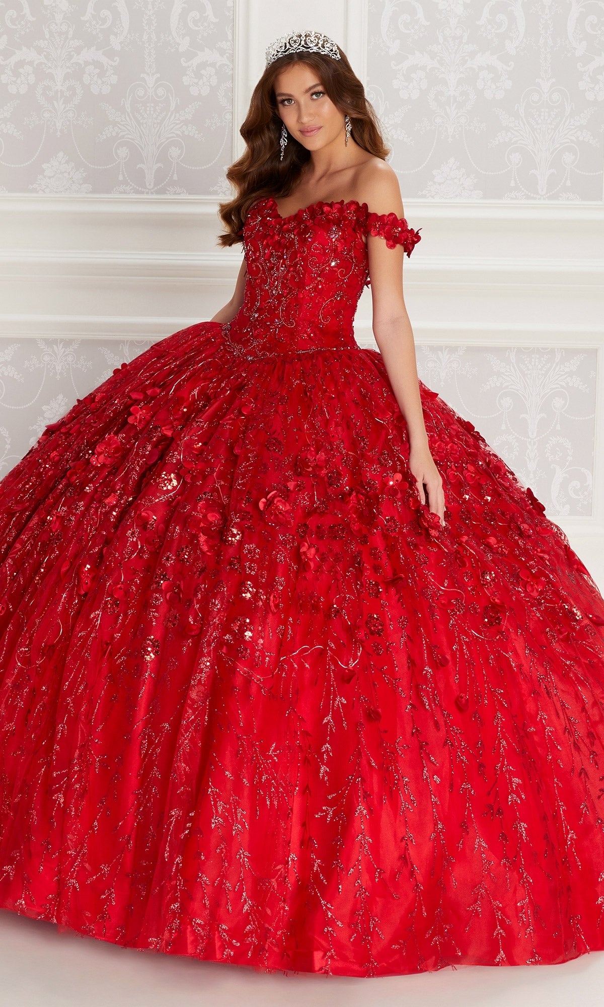 A woman in the Flower Quince Dress, Princesa by Ariana Vara PR22036, featuring a jeweled tiara and elaborate off-the-shoulder red gown with intricate embroidery, stands against a white ornate wall.